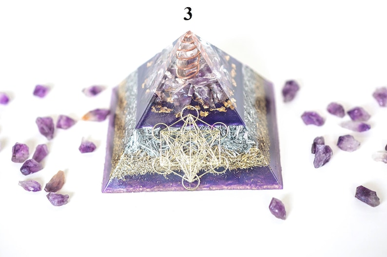May include: A purple and silver pyramid-shaped orgone energy generator with a copper coil at the top. The pyramid features a Metatron's Cube design and is surrounded by amethyst crystals. The number "3" is visible at the top.