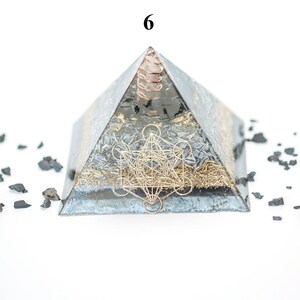 May include: A pyramid-shaped orgone energy generator with a metallic finish, featuring a gold-colored geometric design. The pyramid is surrounded by small, dark-colored stones. The number "6" is visible at the top.