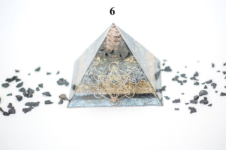 May include: A pyramid-shaped orgone energy generator with a metallic finish, featuring a gold-colored geometric design. The pyramid is surrounded by small, dark-colored stones. The number "6" is visible at the top.