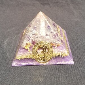 May include: A purple amethyst crystal pyramid with a gold Om symbol on the front. The pyramid is made of clear quartz crystals and has a gold trim.