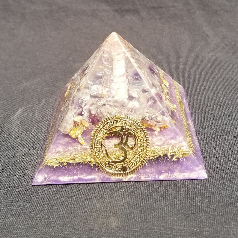 May include: A purple amethyst crystal pyramid with a gold Om symbol on the front. The pyramid is made of clear quartz crystals and has a gold trim.