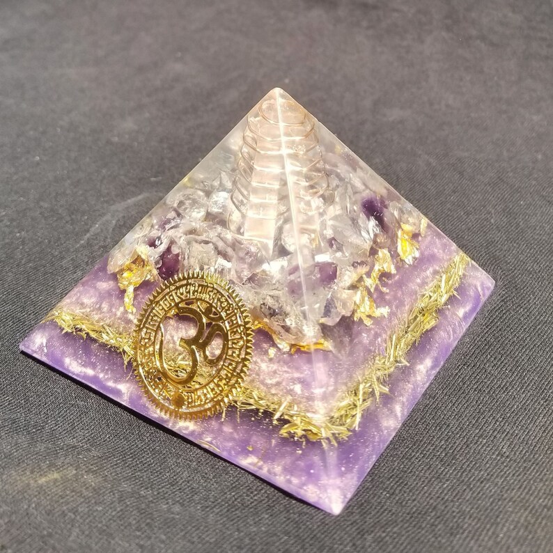 May include: A purple amethyst crystal pyramid with a gold Om symbol and gold flecks. The pyramid has a clear quartz crystal point in the center.