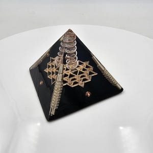 May include: A black pyramid-shaped decorative object with a geometric gold design and a clear crystal at the top. The pyramid has a metallic coil and a star-like pattern. Small copper-colored accents are visible on the sides.