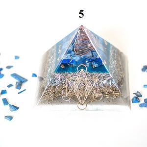 May include: A clear pyramid-shaped orgone energy generator with blue and silver accents. The pyramid features a copper coil at the apex, blue stone inlays, and a silver metal geometric design. Blue stone fragments surround the pyramid.