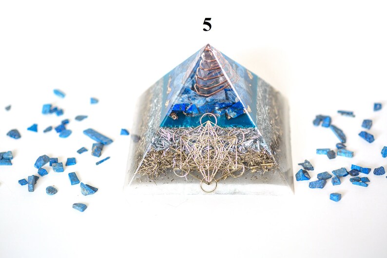 May include: A clear pyramid-shaped orgone energy generator with blue and silver accents. The pyramid features a copper coil at the apex, blue stone inlays, and a silver metal geometric design. Blue stone fragments surround the pyramid.