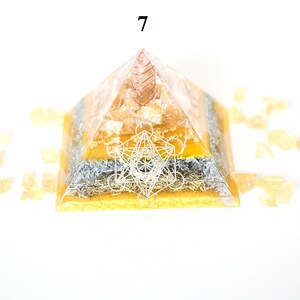 May include: A layered pyramid-shaped object with a geometric design on the front. The pyramid is primarily orange and yellow, with a silver-colored layer. The number "7" is visible at the top. Small, light orange crystals surround the pyramid.