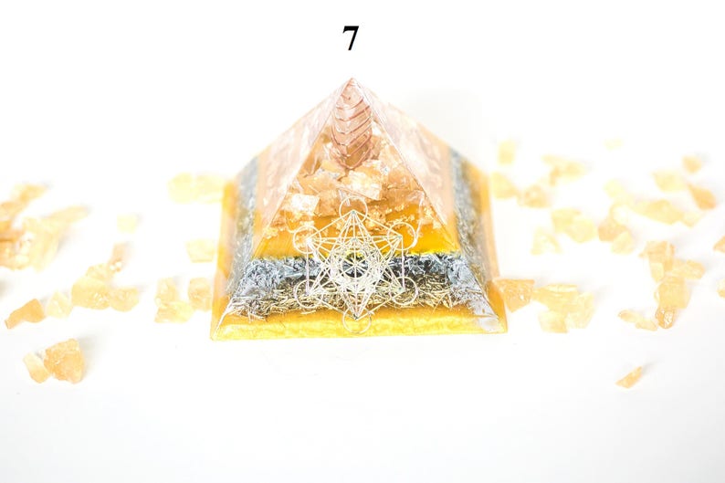 May include: A layered pyramid-shaped object with a geometric design on the front. The pyramid is primarily orange and yellow, with a silver-colored layer. The number "7" is visible at the top. Small, light orange crystals surround the pyramid.
