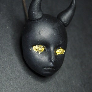 Op de afbeelding: Zwarte ketting met een klein, gedetailleerd gezicht en hoorns. Het gezicht heeft gouden glitterogen.