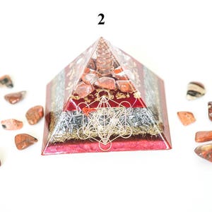 May include: A red and clear pyramid-shaped orgone energy generator with a gold Metatron's Cube design. The pyramid is surrounded by small, polished red and brown stones. The number "2" is visible at the top of the pyramid.