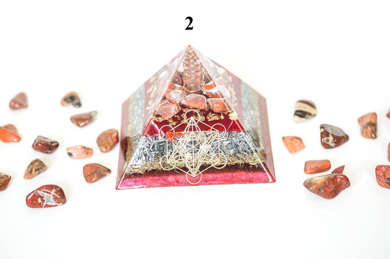 May include: A red and clear pyramid-shaped orgone energy generator with a gold Metatron's Cube design. The pyramid is surrounded by small, polished red and brown stones. The number "2" is visible at the top of the pyramid.