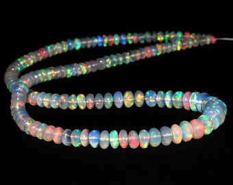 May include: Strand of opal beads with a rainbow iridescence. The beads are round and vary in size, strung on a thin, clear cord against a black background. The opals display flashes of red, green, blue, and yellow.