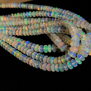 May include: Strands of iridescent opal beads. The beads are a mix of white, blue, green, and orange, with a faceted cut that catches the light. The beads are strung together, creating a flowing, organic shape. The background is black.