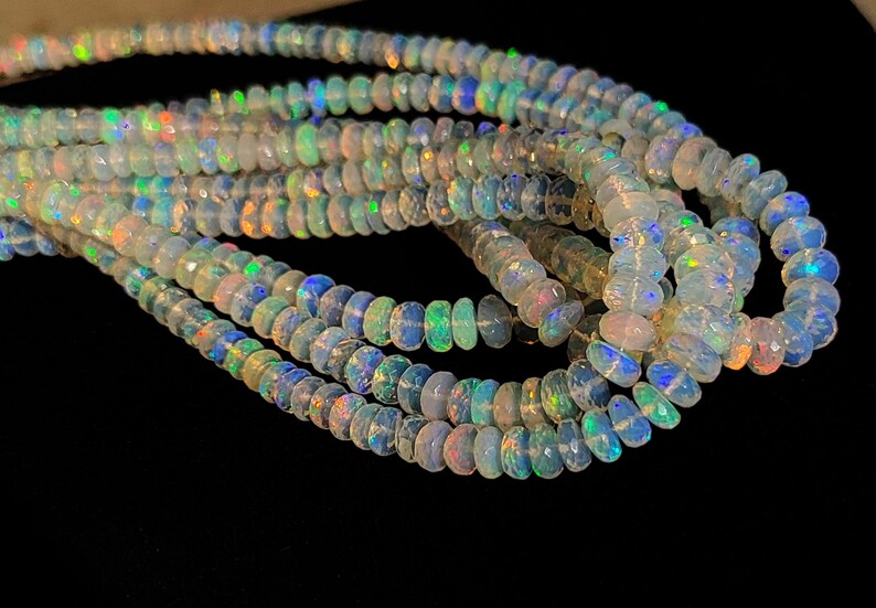 May include: Strands of iridescent opal beads. The beads are a mix of white, blue, green, and orange, with a faceted cut that catches the light. The beads are strung together, creating a flowing, organic shape. The background is black.