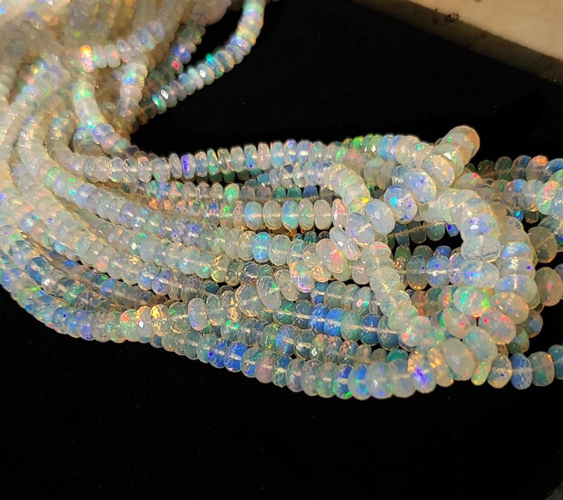 May include: Strands of iridescent opal beads. The beads are a mix of white and translucent colors, with flashes of blue, green, and yellow. The beads are strung together, creating a flowing, textured appearance. The opals are round and faceted.