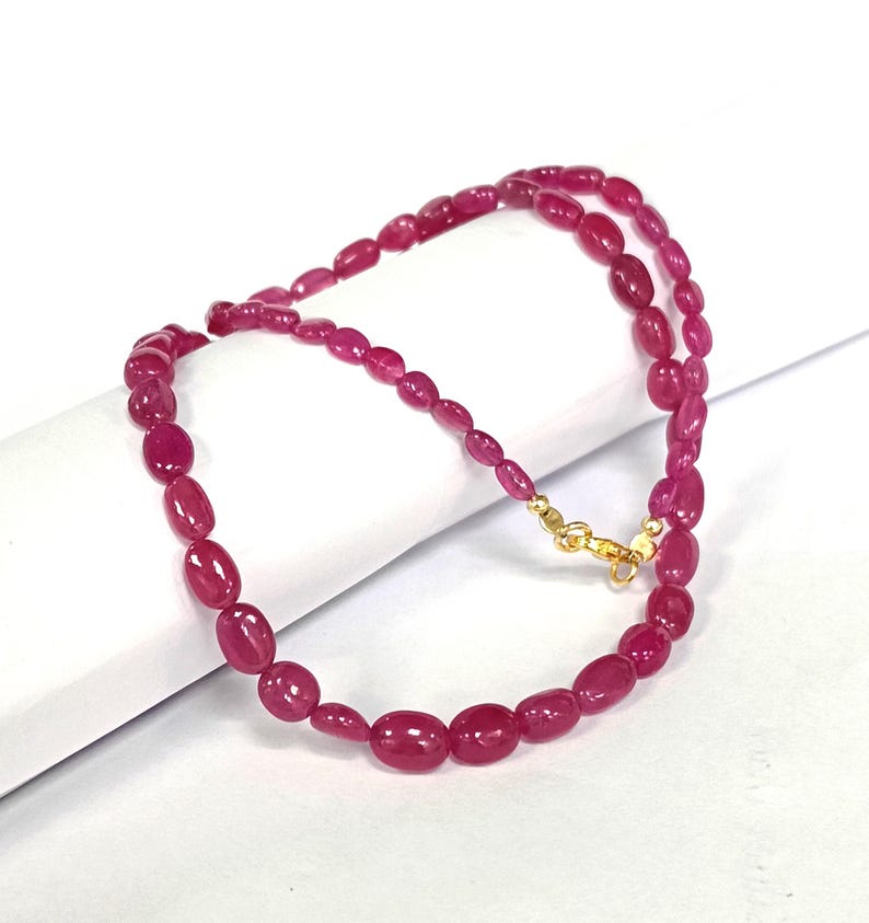 May include: A pink gemstone necklace with oval-shaped beads. The necklace is made of a string of pink gemstones with a gold clasp.