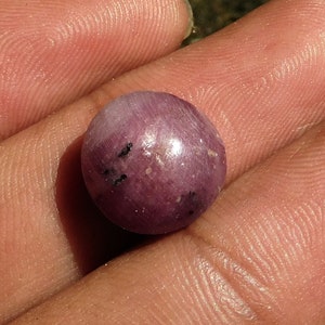 May include: A round, purple gemstone with a shiny, reflective surface. The gemstone has a few small black inclusions.