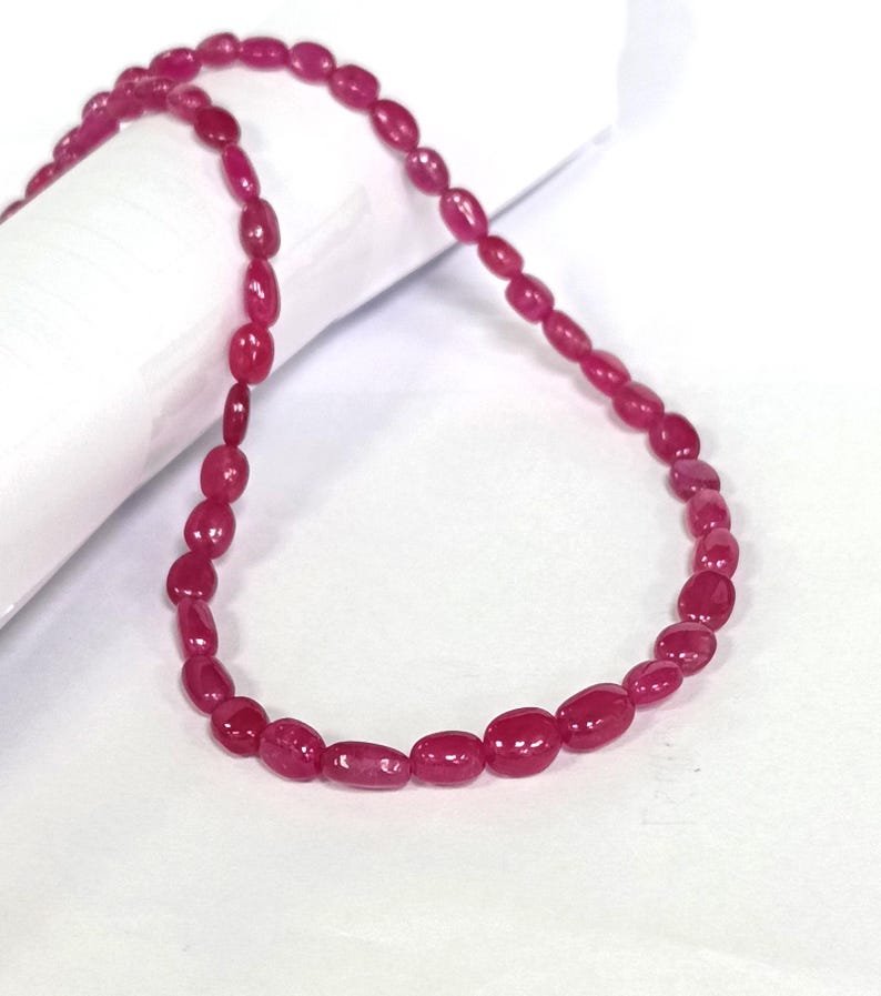 May include: A strand of pink ruby beads, strung together to form a necklace. The beads are oval shaped and have a smooth, polished finish.