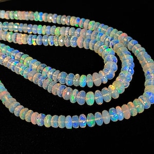 May include: Strands of opalescent gemstone beads. The beads are a mix of white, blue, and iridescent colors, with a faceted cut. The beads are strung together to form necklaces, with a dark background.