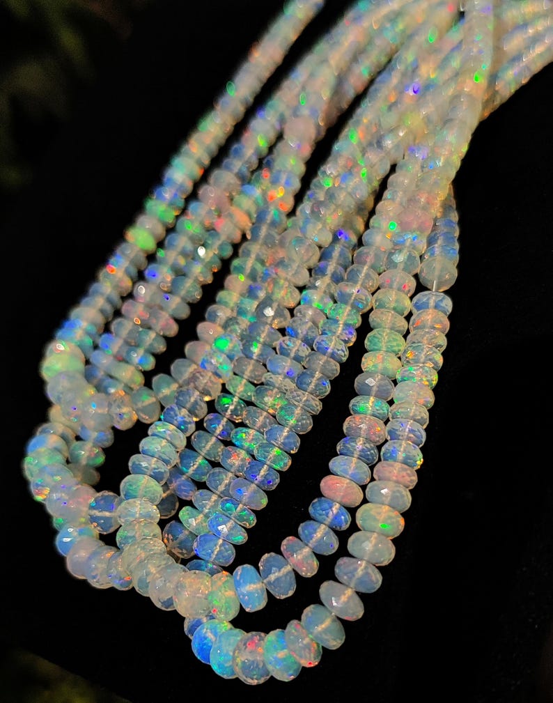 May include: Strands of faceted opal beads with iridescent flashes of color. The beads are a milky white color with hints of blue, green, and pink. The beads are strung together, creating a necklace or bracelet.