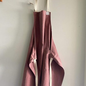May include: A dusty rose coloured apron with white straps. The apron has a single pocket on the front and the text "handmade with love" is printed on a white tag.
