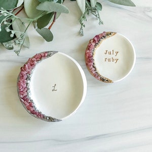 Puede incluir: Dos platos de anillo de cerámica blanca con detalles en rosa y dorado. El plato más grande tiene el texto "July ruby" inscrito. El plato más pequeño tiene la letra "L" inscrita.