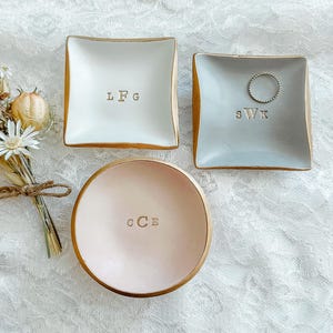 May include: Two square dishes and one round dish with gold-painted edges. The white square dish is stamped with LFG, the light gray square dish features SWK and a silver ring, and the round pink dish is stamped with CCE. A small dried flower bouquet.