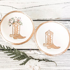 May include: Two white ceramic trinket dishes with gold trim. One dish features a floral design and a gold cowboy boot. The other dish features a gold cowboy boot.
