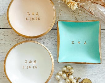 May include: Two round and one square trinket dish with gold-painted edges. The top round dish is pale pink with L heart A and 5.30.25 stamped. The bottom round dish is white with J & S and 2.14.25 stamped. The square dish is teal with K heart A stamped.
