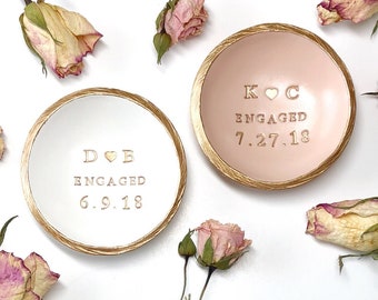 May include: Two round ceramic trinket dishes with gold-painted textured rims. The left dish is white with gold text reading D heart B, ENGAGED, 6.9.18. The right dish is pale pink with gold text reading K heart C, ENGAGED, 7.27.18, surrounded by dried roses.
