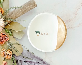May include: White ceramic ring dish with a gold rim and a palm tree design. The dish has the initials "A + R" written on it.