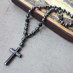 Puede incluir: Un rosario gris oscuro con un colgante de cruz. El rosario presenta cuentas redondas y una cruz en la parte inferior. Las cuentas y la cruz tienen un brillo met&aacute;lico. El rosario se muestra sobre una superficie gris claro.