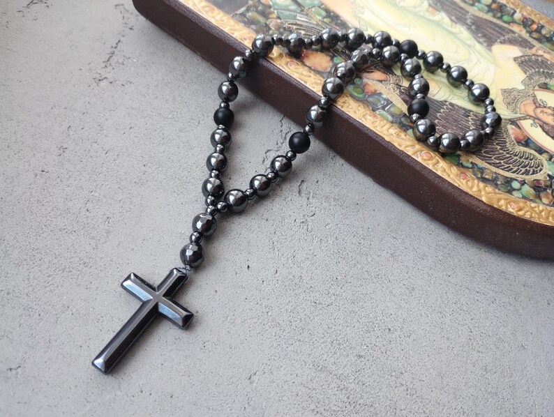 Puede incluir: Un rosario gris oscuro con un colgante de cruz. El rosario presenta cuentas redondas y una cruz en la parte inferior. Las cuentas y la cruz tienen un brillo met&aacute;lico. El rosario se muestra sobre una superficie gris claro.