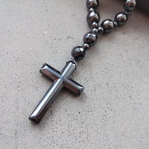 Puede incluir: Un rosario de hematita gris oscuro con un colgante de cruz. La cruz est&aacute; hecha del mismo material que las cuentas y tiene una superficie pulida y reflectante. Las cuentas son redondas y lisas, conectadas por una cadena.