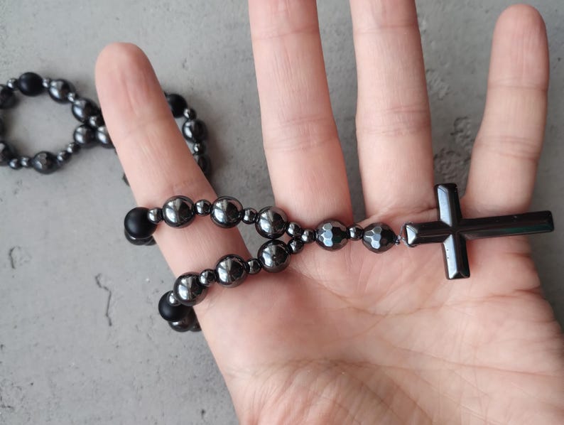 Puede incluir: Un rosario gris oscuro con un colgante de cruz. El rosario presenta cuentas redondeadas y una cruz al final. Las cuentas tienen un brillo met&aacute;lico, y la cruz es de color gris oscuro s&oacute;lido. El rosario se sostiene en una mano.