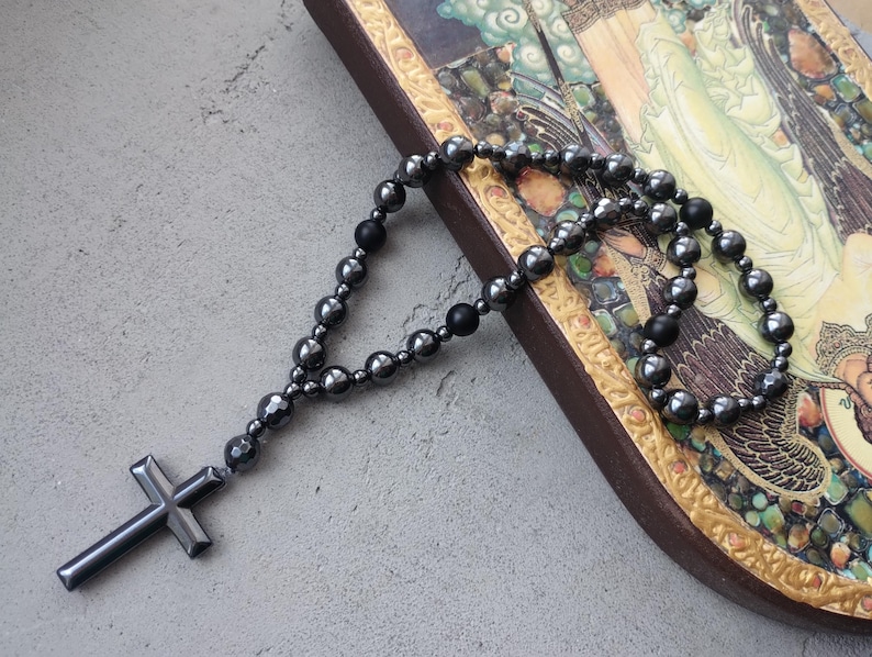 Puede incluir: Un rosario gris oscuro con un colgante de cruz. El rosario presenta cuentas redondeadas y una cruz en la parte inferior. Las cuentas est&aacute;n ensartadas, formando un bucle. El rosario se coloca sobre una superficie decorativa con una imagen.