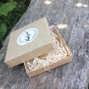 Puede incluir: Una peque&ntilde;a caja de cart&oacute;n marr&oacute;n abierta con papel triturado en su interior, colocada sobre una superficie de madera desgastada. La tapa de la caja est&aacute; parcialmente abierta, revelando el contenido. Una pegatina ovalada blanca en la tapa dice "Thank you!" en escritura a mano.