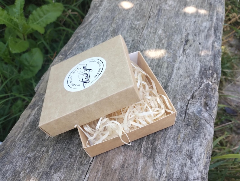 Puede incluir: Una peque&ntilde;a caja de cart&oacute;n marr&oacute;n abierta con papel triturado en su interior, colocada sobre una superficie de madera desgastada. La tapa de la caja est&aacute; parcialmente abierta, revelando el contenido. Una pegatina ovalada blanca en la tapa dice "Thank you!" en escritura a mano.