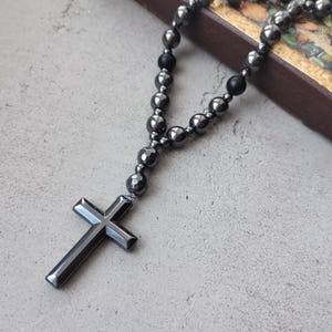Puede incluir: Un rosario gris oscuro con un colgante de cruz. Las cuentas son una mezcla de formas redondas y facetadas, ensartadas para formar un collar. La cruz tiene un dise&ntilde;o simple y pulido. El rosario descansa sobre una superficie gris claro.