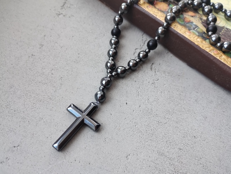 Puede incluir: Un rosario gris oscuro con un colgante de cruz. Las cuentas son una mezcla de formas redondas y facetadas, ensartadas para formar un collar. La cruz tiene un dise&ntilde;o simple y pulido. El rosario descansa sobre una superficie gris claro.