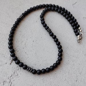 Puede incluir: Un collar de cuentas negras con cierres plateados. Las cuentas son mate y tienen una forma ligeramente irregular.