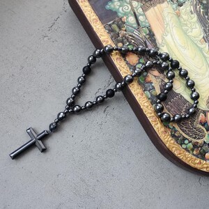 Puede incluir: Un rosario gris oscuro con un colgante de cruz, compuesto por cuentas redondeadas. El rosario descansa sobre un icono religioso decorativo enmarcado con un borde dorado.