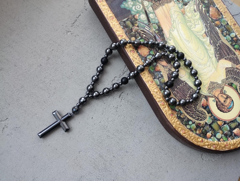 Puede incluir: Un rosario gris oscuro con un colgante de cruz, compuesto por cuentas redondeadas. El rosario descansa sobre un icono religioso decorativo enmarcado con un borde dorado.