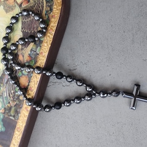 Puede incluir: Un rosario de cuentas negras con un colgante de cruz. Las cuentas redondas y oscuras forman un collar. La cruz tiene un dise&ntilde;o simple y s&oacute;lido. El rosario descansa sobre una superficie con una imagen religiosa.