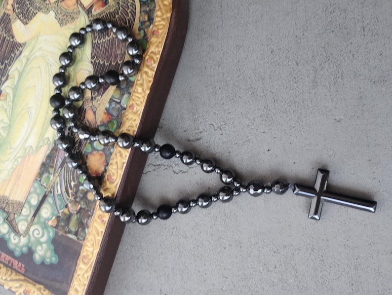 Puede incluir: Un rosario de cuentas negras con un colgante de cruz. Las cuentas redondas y oscuras forman un collar. La cruz tiene un dise&ntilde;o simple y s&oacute;lido. El rosario descansa sobre una superficie con una imagen religiosa.