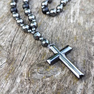 Puede incluir: Un collar con un colgante de cruz plateada y cuentas de color gris oscuro. La cruz est&aacute; hecha de un material met&aacute;lico brillante. Las cuentas son una mezcla de formas redondas y facetadas. El collar descansa sobre una superficie de madera envejecida.
