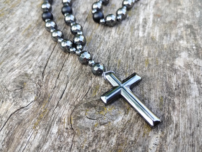 Puede incluir: Un collar con un colgante de cruz plateada y cuentas de color gris oscuro. La cruz est&aacute; hecha de un material met&aacute;lico brillante. Las cuentas son una mezcla de formas redondas y facetadas. El collar descansa sobre una superficie de madera envejecida.