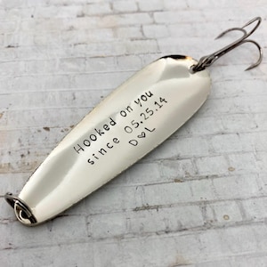 May include: A silver fishing lure with a double hook. The lure has the text "Hooked on you since 05.25.14 D♡L" stamped on the surface. The lure is designed to attract fish.