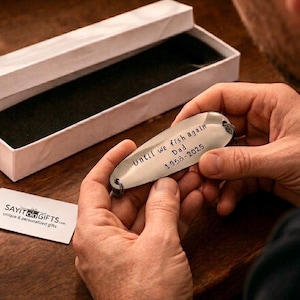 Pu&ograve; includere: Esca da pesca color argento con il testo inciso "Until we fish again Dad 1958-2025". L'esca &egrave; tenuta tra le mani di una persona, vicino a una scatola regalo bianca aperta. &Egrave; visibile un piccolo biglietto bianco con testo.