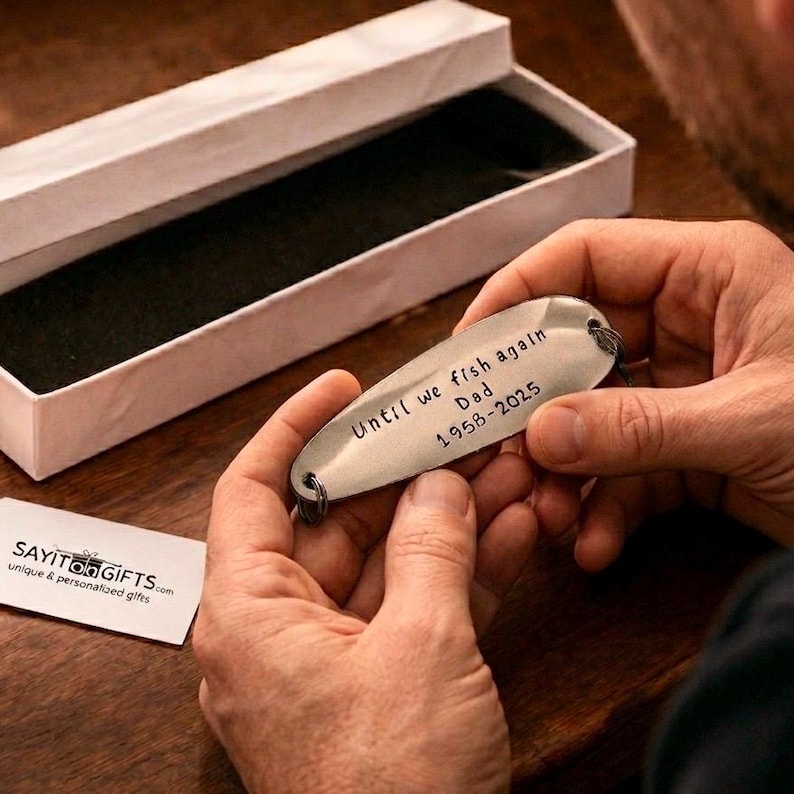Pu&ograve; includere: Esca da pesca color argento con il testo inciso "Until we fish again Dad 1958-2025". L'esca &egrave; tenuta tra le mani di una persona, vicino a una scatola regalo bianca aperta. &Egrave; visibile un piccolo biglietto bianco con testo.