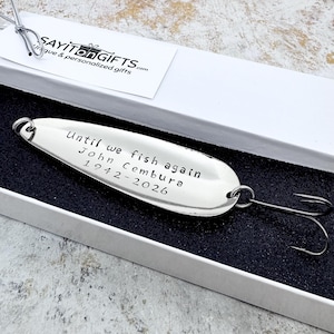 May include: A silver fishing lure with the inscription "Until we fish again John Cembura 1942-2026" is presented in a white gift box. The lure has a hook and is a memorial gift.
